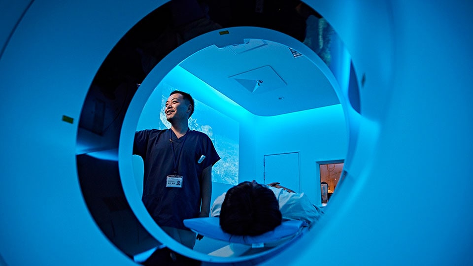 Photo d’un manipulateur qui configure une acquisition sur le statif du scanner, avec le patient sur la table d’examen