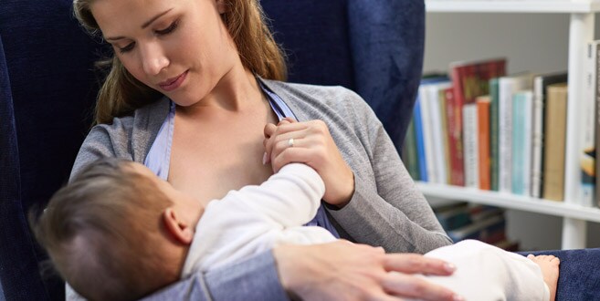 Une maman est assise dans une chaise en velours bleu et regarde son bébé allaiter dans la “position de la berceuse”.