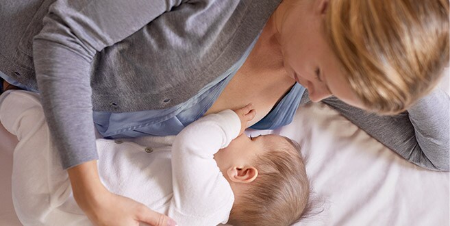 Une maman est allongée sur son côté gauche, sur un lit blanc, berçant et allaitant un bébé dans la position allongée.