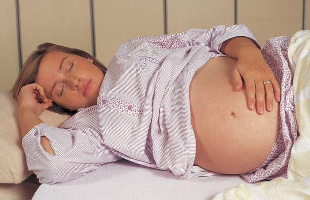Une femme enceinte de plusieurs mois dort sur le côté dans son lit avec une main sur son ventre et l'autre près de son visage.
