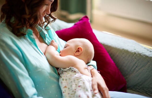 Une mère portant un haut bleu allaite son bébé en position assise sur un canapé gris confortable.