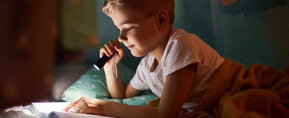 Un garçon lisant un livre avec une lampe torche à la main, allongé sur son lit avec une couverture