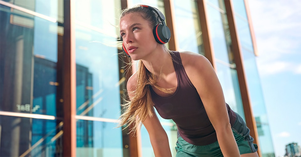Une femme portant un casque Philips A6219 fait une pause lors d'une séance d'entraînement en plein air, debout devant un bâtiment en verre moderne