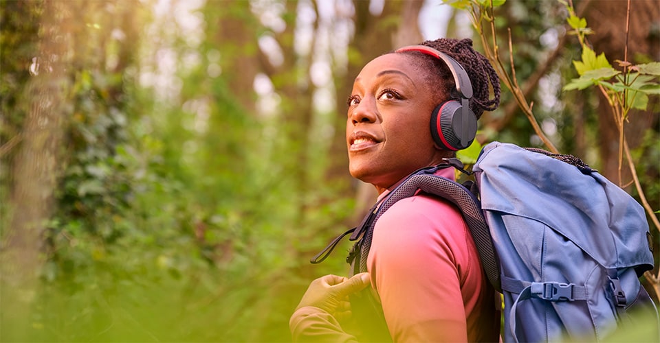 Une femme fait de la randonnée dans une forêt avec un sac à dos bleu, portant un casque Philips A6219 et levant les yeux avec un sourire