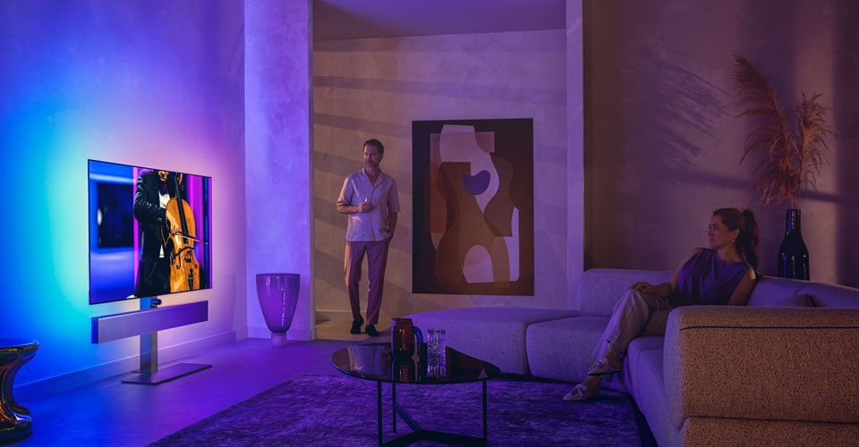Un homme et une femme dans un salon élégant regardent un téléviseur Philips Ambilight diffusant une performance de violoncelle