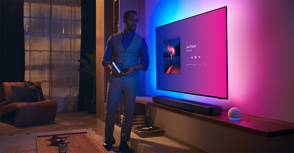 Personne dans un salon moderne près d'un téléviseur Philips Ambilight avec interface musicale et barre de son Fidelio