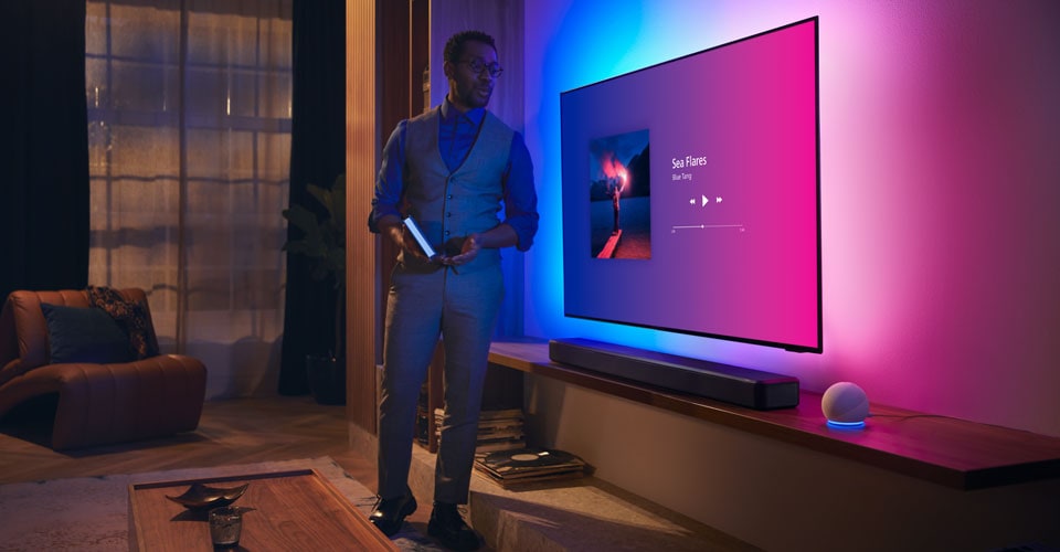 Personne dans un salon moderne près d'un téléviseur Philips Ambilight avec interface musicale et barre de son Fidelio