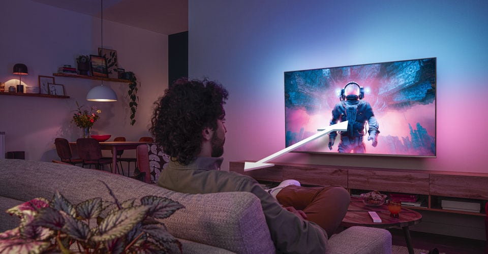 Un homme assis sur un canapé regarde un téléviseur Philips Ambilight affichant une scène d'astronaute colorée, avec une flèche indiquant la distance de visionnage optimale