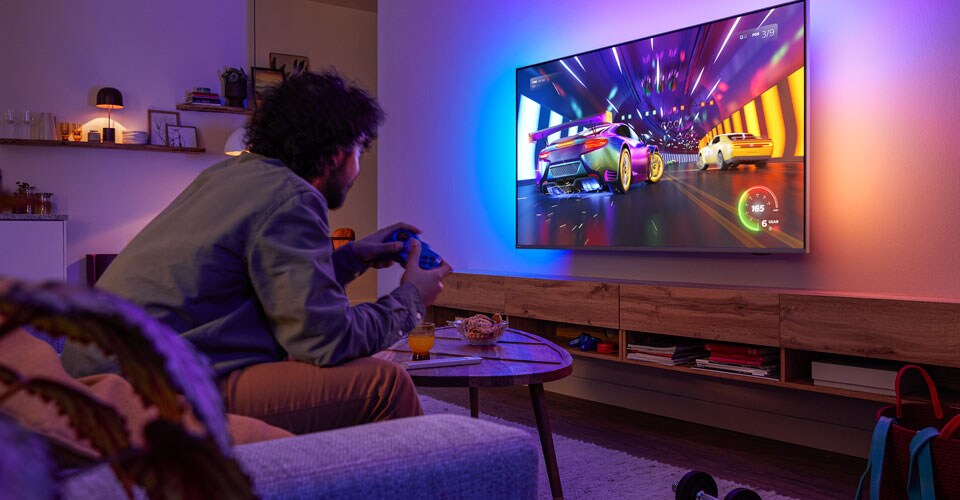 Une personne est assise sur un canapé et joue à un jeu vidéo de course sur un téléviseur Philips Ambilight