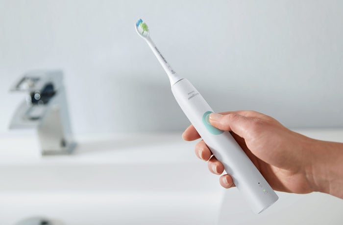 Une main tient une brosse à dents électrique blanche Philips Sonicare en face d’un évier avec le pouce sur le bouton de marche.  