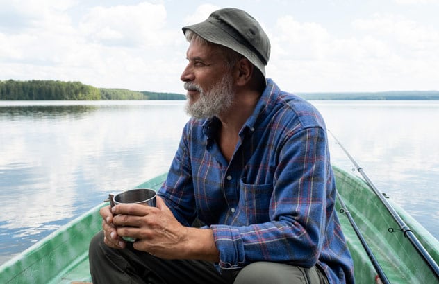 Un homme d’un certain âge portant une barbe grise, un chapeau de pêcheur et une chemise à carreaux bleus est assis sur un bateau et regarde au loin.