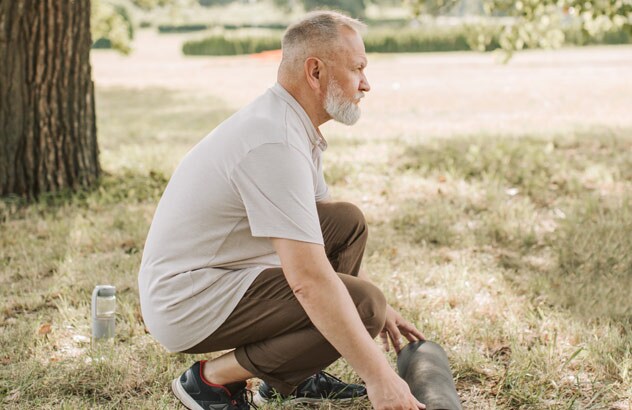 Profil d’un homme avec les cheveux gris et une barbe blanche en train de déplier un tapis de sport dans un pré.