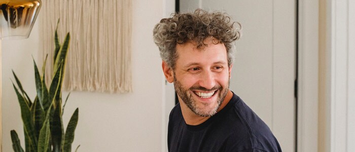 Un homme souriant aux cheveux bouclés et une barbe taillée grise assis devant un piano.