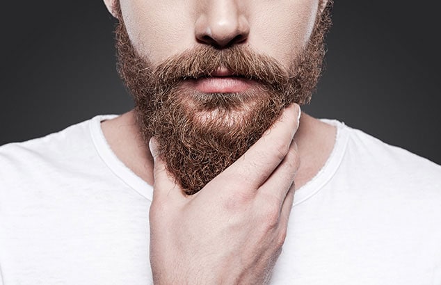 Gros plan d'un homme portant le bas du visage d'un t-shirt, la main dans son épaisse barbe brune.