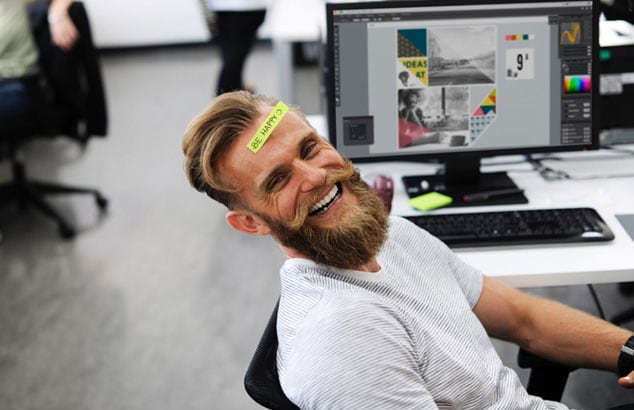 n homme portant une barbe de hipster et un post-it collé sur le front disant “Be Happy” est assis à son bureau.