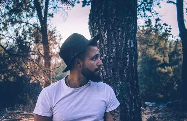 Un homme de profil avec un chapeau et une barbe de hipster se trouve dans la forêt.