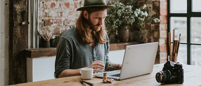 Jeune homme aux cheveux longs et à la barbe pleine assis dans un espace de travail regardant l’ordinateur portable devant lui.