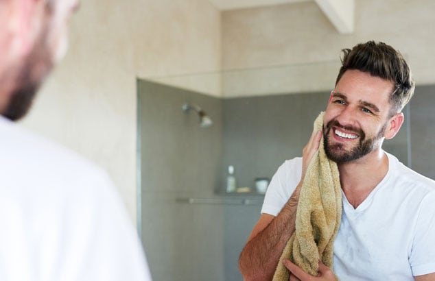 Un homme sourit à son reflet dans le miroir tout en séchant sa barbe épaisse avec une serviette jaune.