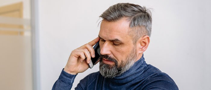 Homme mûr portant une barbe en pointe couleur sel et poivre, habillé en pull à col roulé bleu foncé, regardant sa montre.