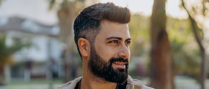 Homme barbu aux cheveux courts foncés portant un dégradé de barbe, souriant dans un jardin verdoyant.