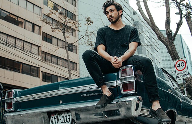 Jeune homme à la barbe et aux cheveux noirs portant des vêtements noirs, assis sur le coffre d'une vieille voiture.