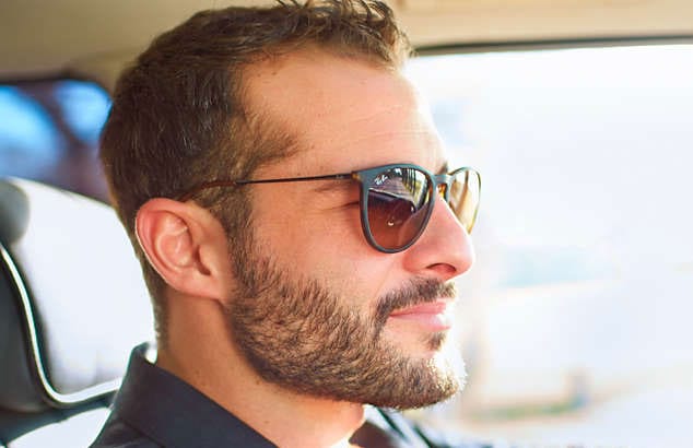 Homme conduisant une voiture avec des lunettes de soleil noires, une barbe épaisse, une moustache brune et une coupe courte.