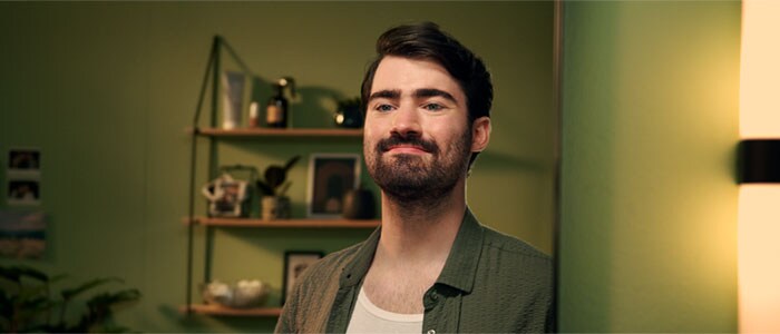 Un homme aux cheveux foncés et une barbe naturellement épaisse sourit dans un miroir dans une pièce au fond vert.