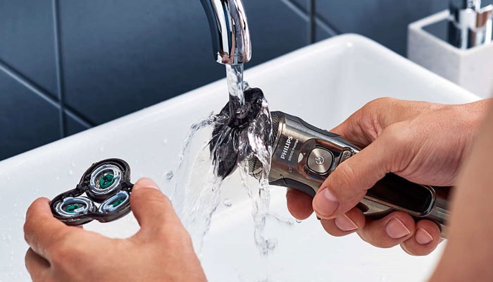 Gros plan d'une personne tenant deux parties d'un rasoir électrique à deux mains sous un robinet d'eau courante.
