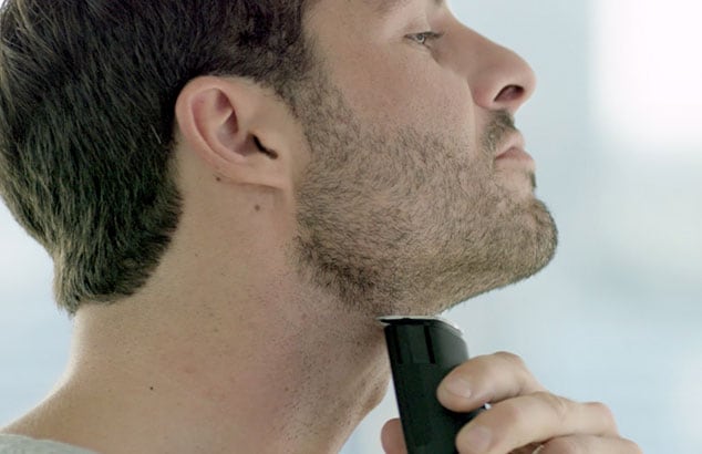 Vue de profil d’un homme portant une barbe courte et se rasant le cou à l’aide d’un rasoir électrique.