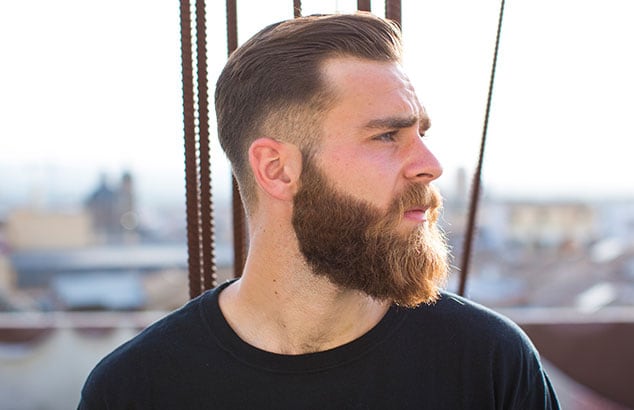 Vue de profil d’un homme brun avec une longue barbe brune, sur le fond d’une ville floutée.
