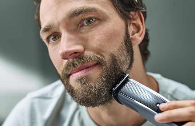 Un homme avec une barbe pleine se rase le visage avec un rasoir électrique argenté et noir.