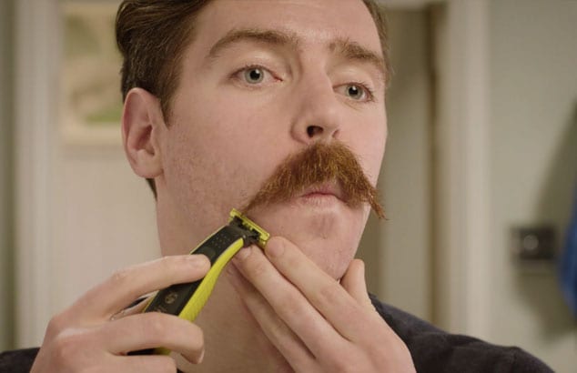 Visage d’un homme avec une moustache qui taille ses pointes à l’aide d’un rasoir électrique.