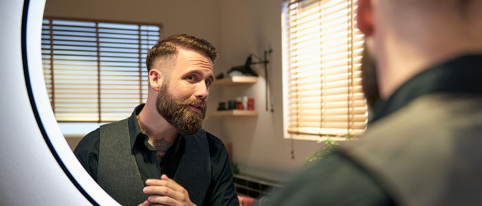 Un homme portant une barbe et un coupe courte dégradé se regarde dans le miroir en souriant après avoir coupé ses cheveux lui-même.