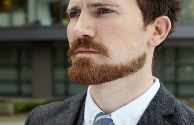 Un homme avec une barbe rousse de menton coupée avec précision dans un costume gris foncé regarde au loin.