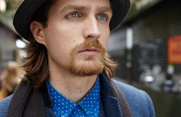 Homme aux cheveux mi-longs et à la barbe henriquatre portant une chemise bleue, une veste de costume et un chapeau.