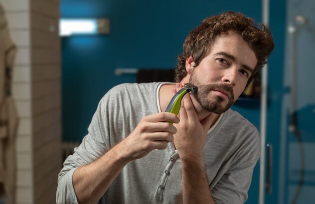 Un homme avec les cheveux bruns se taille sa barbe de trois jours dans sa salle de bain avec son Philips OneBlade.