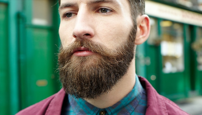 Un homme portant une barbe complète et une moustache se tient debout dans la rue, il porte une chemise à carreaux et une veste bordeaux.